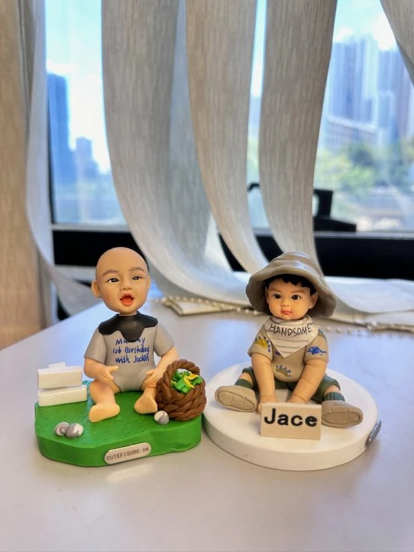 桌面上的兩款小朋友主題人像公仔：左側bb坐在草綠底座，穿淺灰連身衣與深灰圍兜，嘴巴張開微笑，旁有白色木牌造型與小石頭、棕色編織籃內裝綠色葉菜；底座前緣貼有「CUTEFIGURE.HK」小牌。右側bb坐在白色圓底座，戴寬邊卡其漁夫帽、穿米色短袖與短褲，腳著綠色涼鞋，胸前圍巾寫「HANDSOME」，雙腳前放名牌「Jace」。背景為室內窗邊與柔紗簾、遠處高樓景色散景。