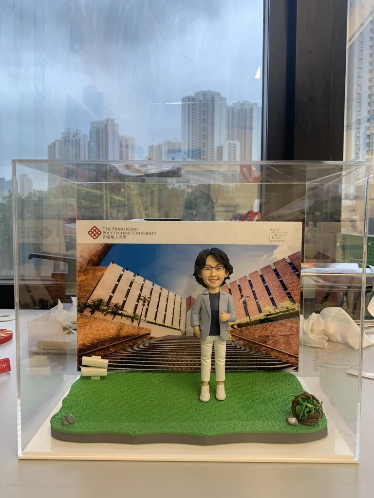透明壓克力展示盒內，一尊短髮戴眼鏡的人像公仔站在綠色草地質感底座上，穿淺灰外套、深色上衣、白色九分褲與白鞋，右手比讚、左手插口袋。背板是一張校園建築階梯的照片，左上角印有「The Hong Kong Polytechnic University／香港理工大學」及紅色結形校徽，右上角標示「College of Professional and ...」等英文字樣。底座右前角有一小叢多肉植物與石頭裝飾；盒外背景透出城市高樓景色。