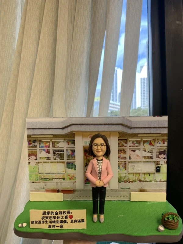 一款老師主題的人像公仔置於校園場景底座：公仔留短髮、戴眼鏡，穿粉色外套、印花上衣與黑色長褲，雙手於身前交握；腳踏灰色地磚，站在綠色草地底座上。背景板為幼稚園/兒童學校門口與窗格，窗內有彩繪與兒童圖案。前方立有中文祝福牌匾，內容祝賀校長退休；右側有小花籃與白色小裝飾物。整體陳列在室內、靠近落地窗與窗簾。