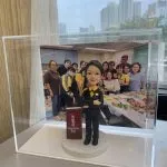 透明壓克力展示盒內，一個穿黑色西裝、黃色襯衫的人像公仔雙手交疊站立，腳下為白色圓形底座；旁邊立著寫有「九龍灣 Kowloon Bay」的紅色站牌造型。展示盒後方放置一張團體合照，十多位穿黃黑制服與便服的人在辦公室/茶水間前合影，桌面擺滿食物與飲品。窗外可見高樓天際線與陰天。