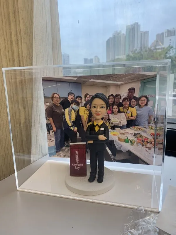 透明壓克力展示盒內，一個穿黑色西裝、黃色襯衫的人像公仔雙手交疊站立，腳下為白色圓形底座；旁邊立著寫有「九龍灣 Kowloon Bay」的紅色站牌造型。展示盒後方放置一張團體合照，十多位穿黃黑制服與便服的人在辦公室/茶水間前合影，桌面擺滿食物與飲品。窗外可見高樓天際線與陰天。