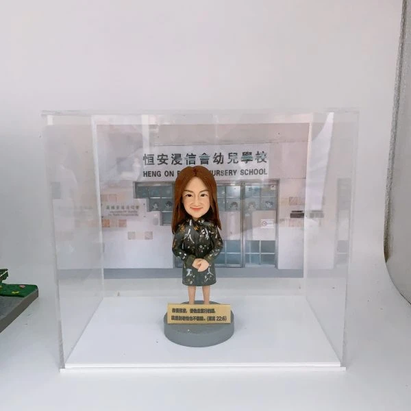 餞別禮物人像公仔，長髮女性，穿着深色碎花裙裝，雙手合十站立於灰色圓形底座，底座刻有文字，背板印有「恒安浸信會幼兒學校」校舍門口，整體收納於透明展示盒中。