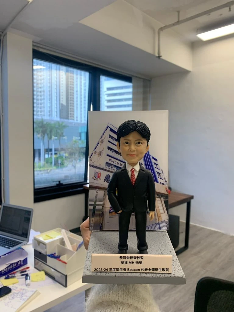 老師禮物人像公仔，人物黑髮，身穿黑色西裝配白色恤衫及紅色領帶，雙手自然放在身側，站立於灰色方形底座，上有金屬銘牌刻有文字，背板印有學校名稱。