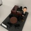 人像公仔辦公室場景：短髮男性公仔穿深色西裝外套與長褲、內搭淺藍襯衫，腳穿棕色皮靴，側身倚靠木質主管桌，雙手自然交疊。桌上配置筆電、螢幕支架、電話座與文具收納；一旁有黑色辦公椅與圓形會議桌，地面為帶有彩色線條的地毯紋理。整體呈現主管/經理級專業辦公氛圍的人像公仔擺設。