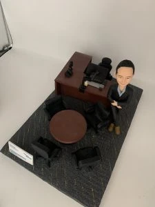 人像公仔辦公室場景：短髮男性公仔穿深色西裝外套與長褲、內搭淺藍襯衫，腳穿棕色皮靴，側身倚靠木質主管桌，雙手自然交疊。桌上配置筆電、螢幕支架、電話座與文具收納；一旁有黑色辦公椅與圓形會議桌，地面為帶有彩色線條的地毯紋理。整體呈現主管/經理級專業辦公氛圍的人像公仔擺設。