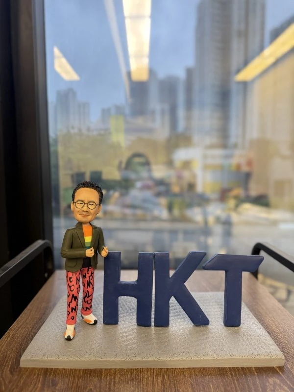 香港電訊(HKT)特色人像公仔，高層人物造型，戴黑框眼鏡，着綠色外套配彩色橙色上衣，紅色長褲印有黑色字樣，腳踏白鞋，站在灰色石紋底座，身旁有深藍色「HKT」立體字母，適合作為高層禮物或公司紀念品。