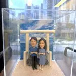 透明展示盒中的情侶人像公仔場景：米色砂地質感底座上，一座淺米色鳥居立於中央，後方為岩石與藍色海景、天空雲紋背板。左側角色穿藍色上衣與黑褲，右側角色穿藍灰色綁帶襯衫裙與白鞋，兩人面向前方、在鳥居前牽著彼此的手。展示盒置於木桌旁，窗外可見高樓與街景倒影。