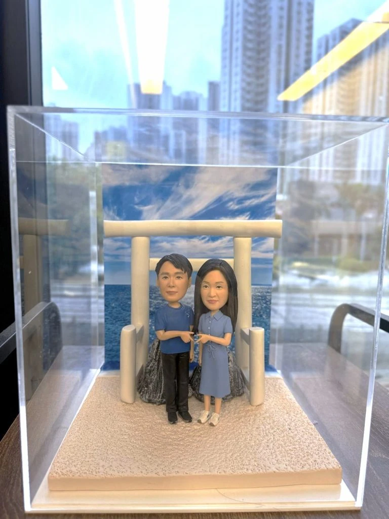 透明展示盒中的情侶人像公仔場景:米色砂地質感底座上,一座淺米色鳥居立於中央,後方為岩石與藍色海景、天空雲紋背板。左側角色穿藍色上衣與黑褲,右側角色穿藍灰色綁帶襯衫裙與白鞋,兩人面向前方、在鳥居前牽著彼此的手。展示盒置於木桌旁,窗外可見高樓與街景倒影。