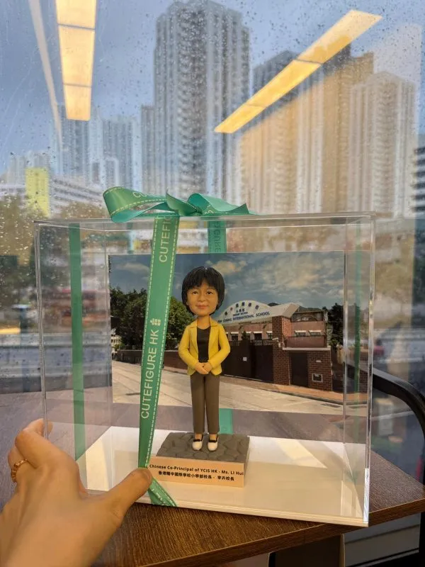 透明展示盒內的人像公仔站立於方形底座上：角色穿黃色西裝外套、黑色上衣與灰色長褲，雙手交疊於身前；身後放置學校外牆與校門的相片式背景。展示盒以綠色CUTEFIGURE HK緞帶綁結，前方名牌印有中英文職銜與名稱字樣；盒子置於木桌上，窗外雨滴與高樓景象映入玻璃。