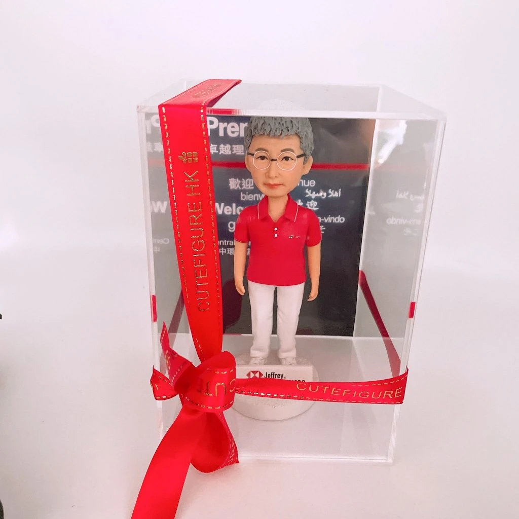 一個透明壓克力展示盒以紅色緞帶環繞並綁成蝴蝶結，盒內站立著銀行職員主題的人像公仔：短髮、配戴圓框眼鏡，穿紅色翻領短袖上衣與白色長褲、白鞋，腳下為白色圓形台座，台座前緣有銀行標誌與姓名標籤。展示盒後方的背板印有多語言「Welcome」字樣與深色背景，整體置於白色桌面上，畫面簡潔、禮盒感十足。