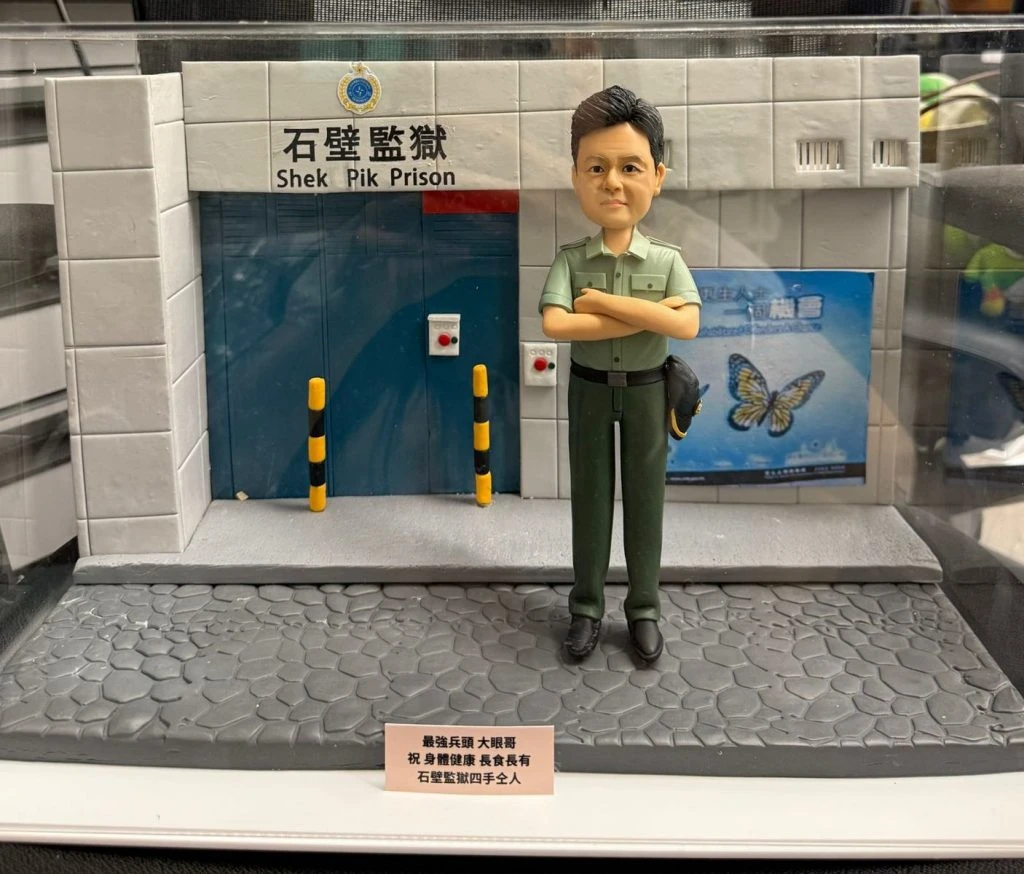 透明展示盒中的客製模型：前景是石紋灰色地坪與小銘牌，中央站著一位雙臂交叉的男性人像公仔，短髮、穿淺綠制服上衣與深綠長褲、黑皮鞋，腰側掛帽。背後是石壁監獄入口的立體建築場景：白灰色方格牆、藍色金屬大門、上方有中英文「石壁監獄 Shek Pik Prison」招牌與徽章，門旁設有兩根黃黑相間防撞柱與緊急按鈕盒，右側牆面貼有藍底蝴蝶宣導海報，整體細節清晰，呈現退休紀念主題。