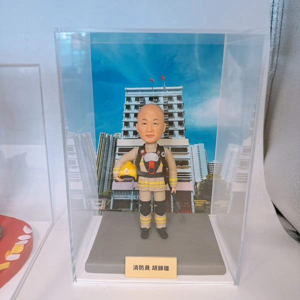 透明展示盒內擺放一個穿卡其黃色消防制服的人像公仔，胸前有呼吸器造型配件，手臂抱著黃色安全帽，腳穿黑色靴子。公仔站在灰色底座上，前方金色銘牌刻有「消防員」與姓名字樣。背景照片是藍天之下的多層白色建築與城市高樓，襯托消防主題氛圍。