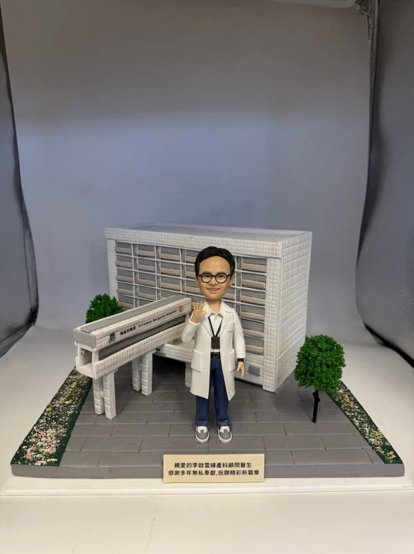 多張照片展示一組醫療主題的人像公仔場景：穿白袍、戴眼鏡的公仔站在灰色地磚平台上，頸掛證件、右手舉起示意。背後是白色方格磚紋的大型建築模型，立面有橫向百葉結構，前方連接一段高架走道與入口雨棚；平台兩側種有綠色圓頂小樹與鋪滿花瓣的綠化帶。前方小銘牌以中文寫上致謝字樣。部分照片顯示整套作品收納於透明展示盒，綁有綠色緞帶。