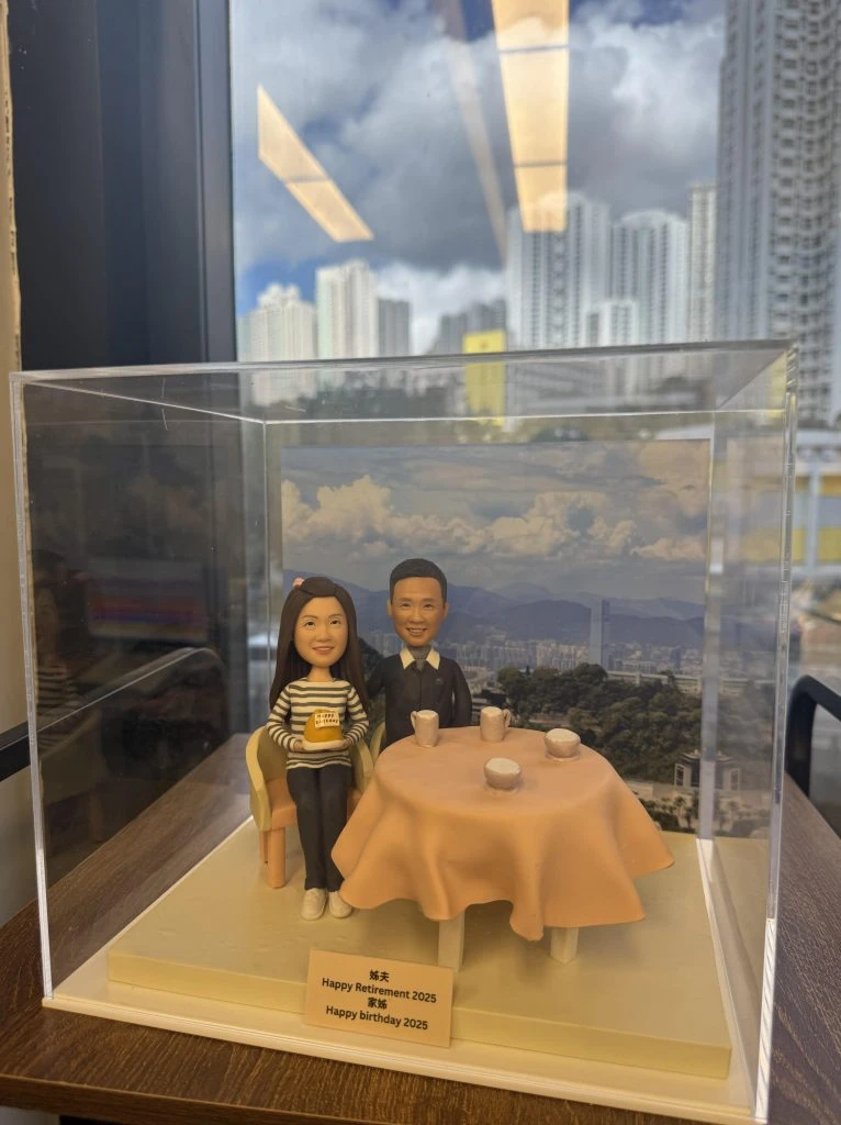 透明壓克力展示盒中的雙人退休人像公仔：左側角色穿條紋上衣、黑褲與白鞋，雙手捧杯入席；右側角色著深色西裝，並肩坐在圓桌旁。桌面覆粉橘色桌布，擺放三只茶杯與一只小盅，呈現酒樓用茶氣氛。前方小銘牌寫有「Happy Retirement 2025」與「Happy birthday 2025」。背板為城市高樓與遠山景致，整體置於木質檯面。