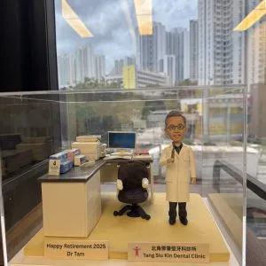 一個放在透明壓克力盒內的牙科主題場景：穿白色醫師袍的牙醫人像公仔站在米黃色底座上，旁邊是深色診療椅與白色診療檯，檯面擺有文件盒、口罩盒與工具罐。右前角的金色銘牌寫著「Happy Retirement 2025 Dr Tam」，側邊垂直銘牌標示「Tang Siu Kin Dental Clinic 北區鄧兆堅牙科診所」。背景透過窗景可見高樓與藍天。