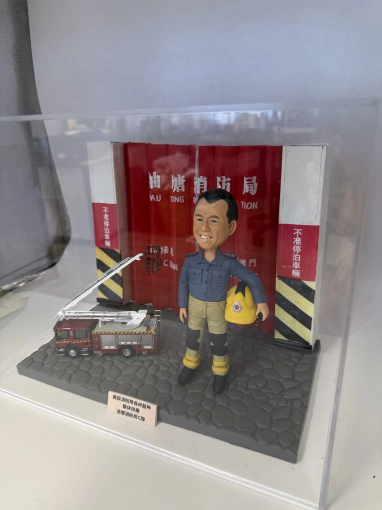 透明展示盒內的人像公仔場景：一位消防主題公仔站在灰色六角石紋台座上，身穿深藍上衣與卡其色防護褲、黑靴與護膝，雙腿綁有黃色反光帶；左臂抱著黃色消防頭盔。背景是紅色消防局摺疊車庫門與黑黃斜紋防撞柱，門上可見白色「消防局／FIRE STATION」字樣。公仔右前方擺放一輛紅色雲梯消防車模型，台座前角有淺色小銘牌，上印黑字祝賀訊息。整體置於透明壓克力盒中。
