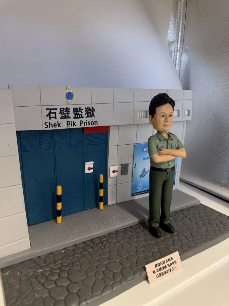 透明展示盒中的客製模型：前景是石紋灰色地坪與小銘牌，中央站著一位雙臂交叉的男性人像公仔，短髮、穿淺綠制服上衣與深綠長褲、黑皮鞋，腰側掛帽。背後是石壁監獄入口的立體建築場景：白灰色方格牆、藍色金屬大門、上方有中英文「石壁監獄 Shek Pik Prison」招牌與徽章，門旁設有兩根黃黑相間防撞柱與緊急按鈕盒，右側牆面貼有藍底蝴蝶宣導海報，整體細節清晰，呈現退休紀念主題。