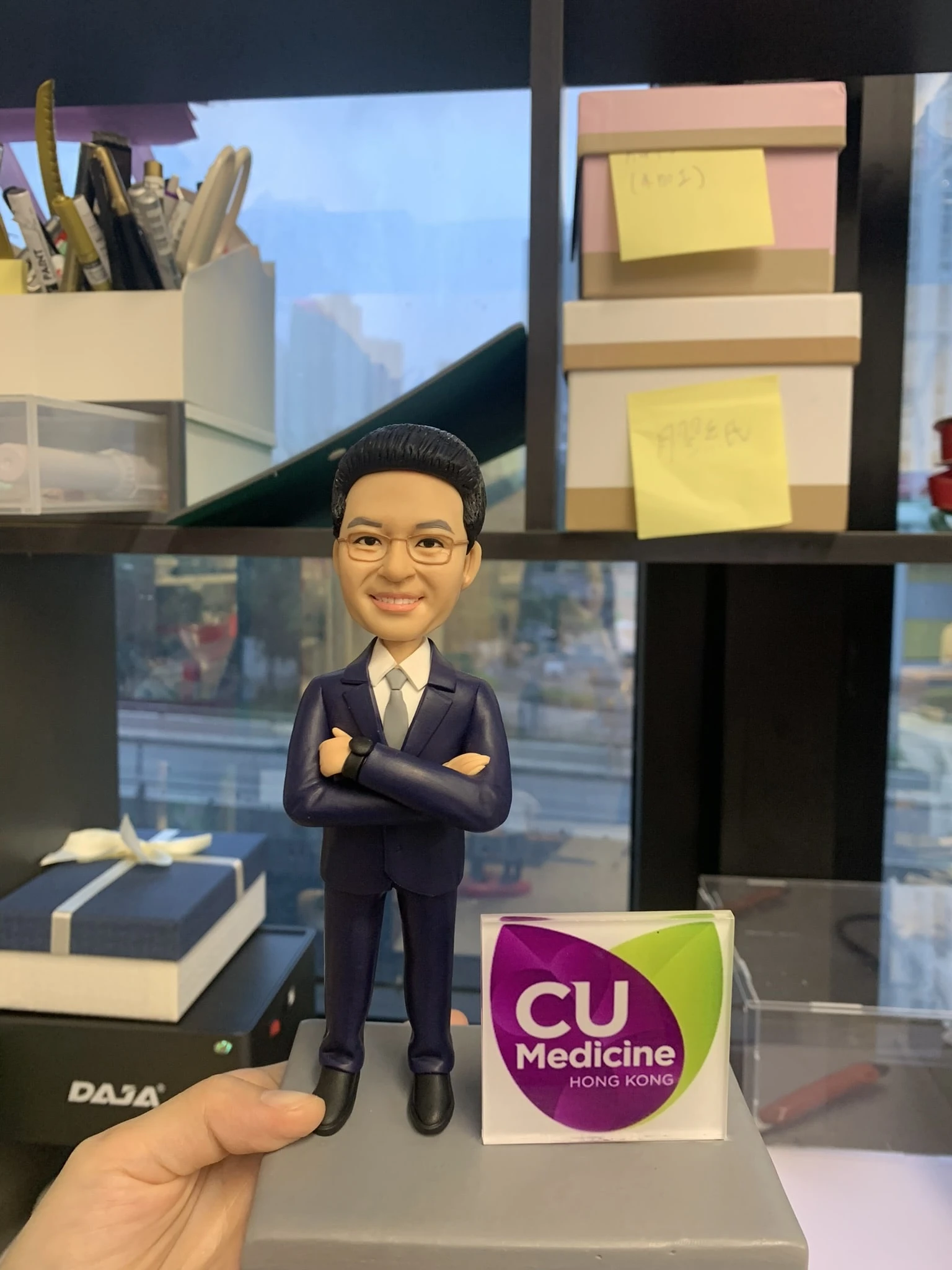 (客製化「香港中文大學醫學院」專業形象公仔 Q版專業男士公仔（身穿深藍色修身西裝套裝，搭配白色襯衫與淺灰色領帶，展現沉穩幹練的菁英氣質。雙手自信交叉於胸前，左手腕佩戴黑色手錶，細節刻畫入微。面部特徵捕捉精準，戴著細框眼鏡，髮型整潔俐落，面露自信且親切的微笑，散發出專業人士的可信賴感） 專屬品牌標誌底座（灰色方形底座，穩重踏實。右側立有顯眼的透明壓克力牌，印有香港中文大學醫學院（CU Medicine Hong Kong）的官方標誌，紫綠配色的Logo清晰可見，象徵著學術權威與醫療專業的身份認同） Q版寫實風、商務專業、醫學菁英、CUHK校友、職人手作、形象塑造 醫師禮物、醫學院畢業紀念、診所開業賀禮、升遷榮退紀念、辦公室專業擺飾)