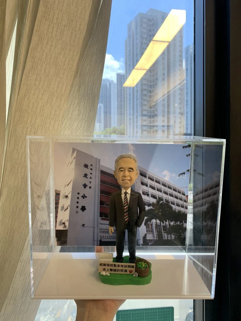 一個放在透明展示盒內的桌面人像公仔。公仔為短灰髮、微笑表情的男性形象,穿深色西裝與白襯衫、紅黑條紋領帶,右手自然下垂、左手插袋,站姿端正。腳下是綠色草地造型底座,左前方有籐籃與綠植裝飾,前側白色牌子印有中文感謝與祝福字樣。背板是一棟多層校舍的照片,可見立面、窗戶與棕櫚樹,牆上有中文校名。展示盒由手掌托起,背景是室內窗邊、米色窗簾與窗外高樓、藍天白雲。