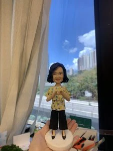 客製化Q版女士公仔——比心手勢休閒造型 手工人物公仔細節 （圖片展示了一尊客製化的Q版女性人物黏土公仔，主角留著及肩的黑色微捲短髮（Bob頭），氣質優雅，面帶溫柔微笑。她身穿一件黃色底配白色與綠色碎花圖案的短袖上衣，下身搭配黑色寬腳褲（Culottes），腳踩黑色尖頭高跟鞋，造型時尚且休閒。公仔最獨特的動作是雙手在胸前比出「愛心」手勢，展現出親切、充滿愛意與關懷的形象。公仔站立在一個簡約的白色圓形底座上。） 環境背景與光影 （公仔由一隻手托起展示，背景位於室內窗邊。左側有垂下的米白色窗簾，窗外是大片蔚藍的天空、白雲以及遠處的城市樓景與綠樹，還有下方的高架道路。光線充足且自然，明亮的日光讓公仔的膚色與衣著色彩顯得格外清晰自然。下方工作檯上可見橙色美工刀、畫筆等製作工具，顯示這是在工作室內完成後的實拍記錄。） 軟陶公仔、真人公仔訂製、Q版人像、女士禮物、比心手勢、休閒造型、手工禮物、黃色碎花衫、寬腳褲穿搭、生日禮物 表達愛意、溫馨紀念、感謝心意、母親節禮物、週年紀念、獨一無二、辦公室擺設、幸福時刻