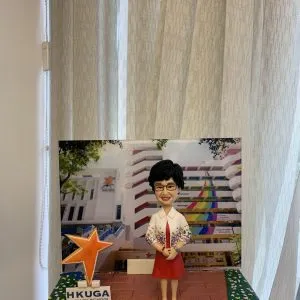 手托一座小型桌面場景模型：中央是一位短髮、戴眼鏡的人像公仔，穿白色外套搭配彩色花卉印花袖口與紅色連身裙，雙手於腹前交握站立。背景立牌印有一所學校建築照片，立面帶有紅色水平線與彩虹色斜紋裝飾，四周樹葉遮蔭。左前方立著一顆橙色星形標誌與「HKUGA PRIMARY SCHOOL」字樣的小牌。底座為紅棕色磚紋地面，兩側綠色帶狀區域點綴白色小花；前方金屬銘牌寫著「HKUGA Primary School Principal Wong Kwai Ling」。