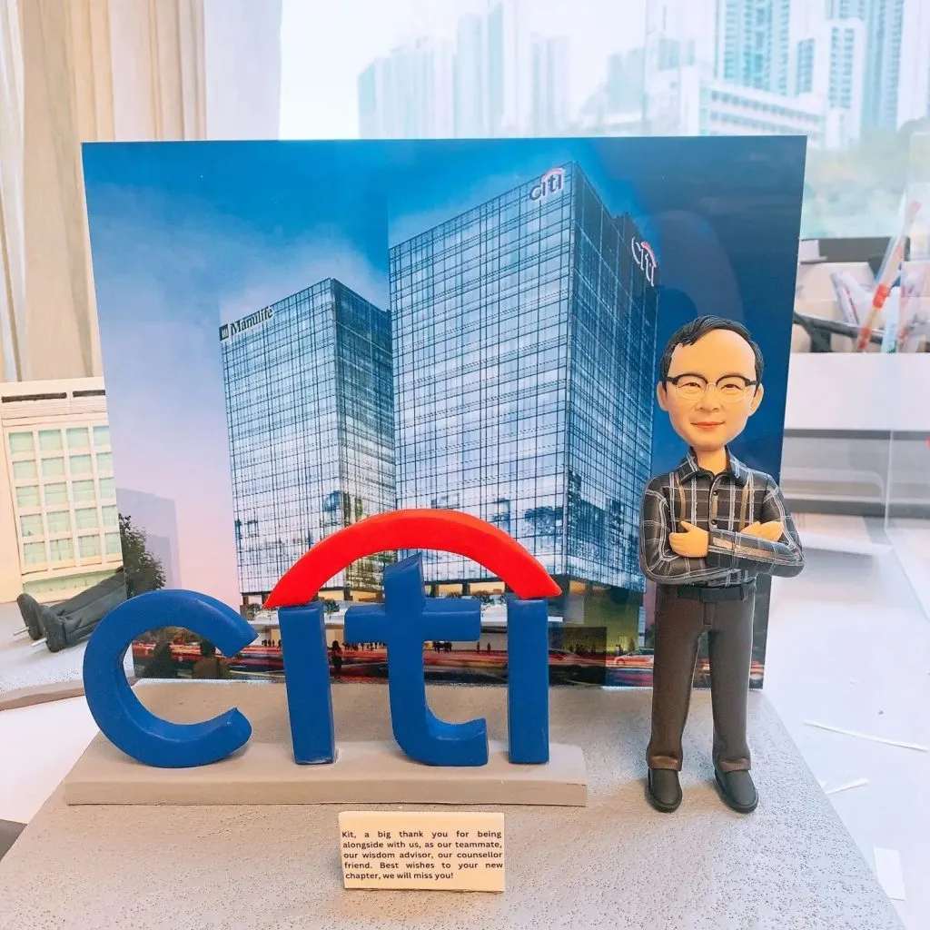 一組桌面擺設：前景是藍色「citi」立體字母與紅色弧形拱，置於灰色台座上；右側站著一個雙臂交叉的人像公仔，短髮、戴眼鏡，穿黑灰格紋襯衫與深色長褲、黑鞋。台座前方有一塊英文訊息小銘牌。背後立著一張高樓大廈照片作為背景板，玻璃帷幕上可見品牌標記與城市天際線；周圍是工作枱與文具雜物。