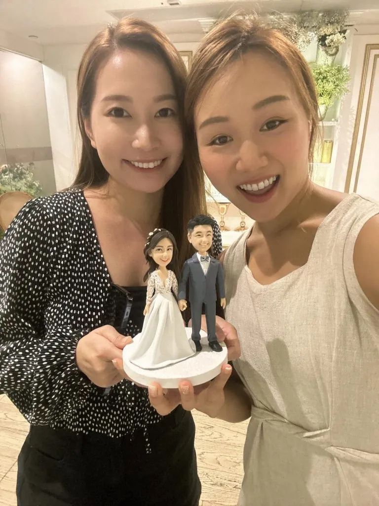 兩位女性在明亮室內對鏡頭微笑合影，雙手托著一座圓形白色台座的人像公仔。公仔為婚禮主題雙人組：左側新娘穿白色長裙禮服，裙擺層次與蕾絲袖設計明顯，髮上點綴小花；右側新郎穿深灰西裝、白襯衫與蝴蝶結，黑色皮鞋。背景為淺色牆面與層架，上擺放綠植與裝飾品，地板為淺木色紋理。
