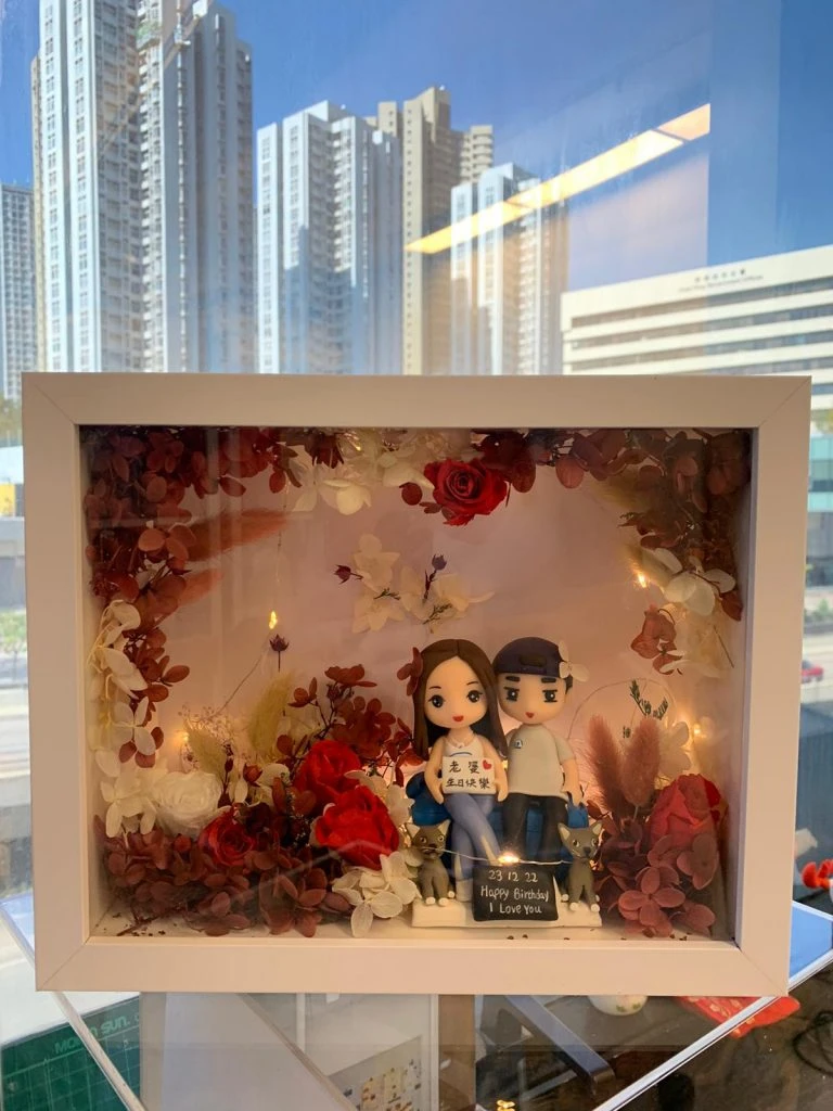白色立體相框中，Q版情侶肩並肩坐著，女孩拿字牌、男孩戴鴨舌帽，腳邊兩隻灰色小貓；相框四周以紅白乾燥花與燈串環繞，底部寫有日期與祝福。