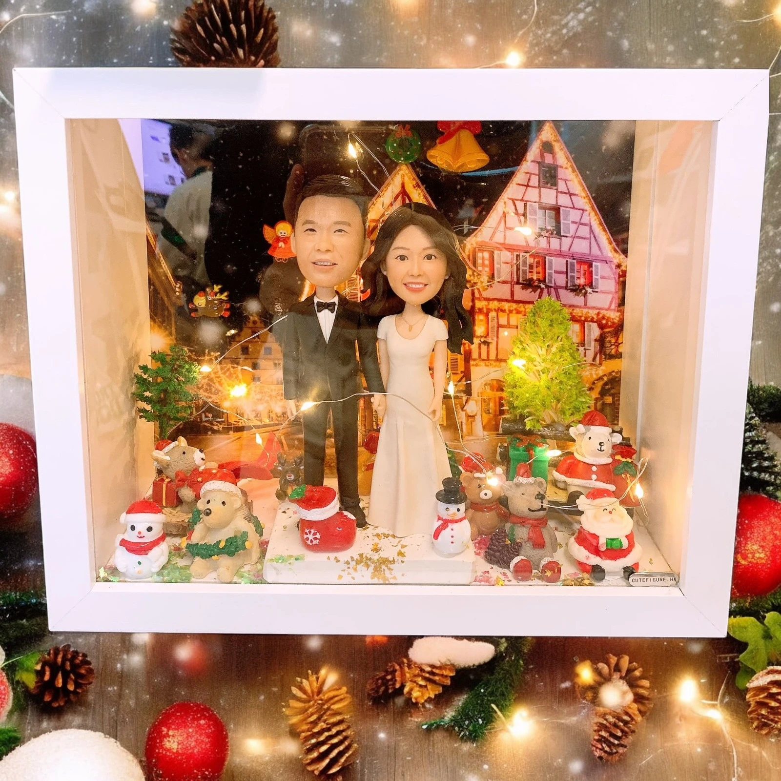 白色相框內的聖誕小鎮場景：身穿禮服與白色洋裝的情侶公仔站在中央，周圍有聖誕樹、雪人、麋鹿與禮物小物，背景為燈火通明的木屋街景，暖白燈串環繞。