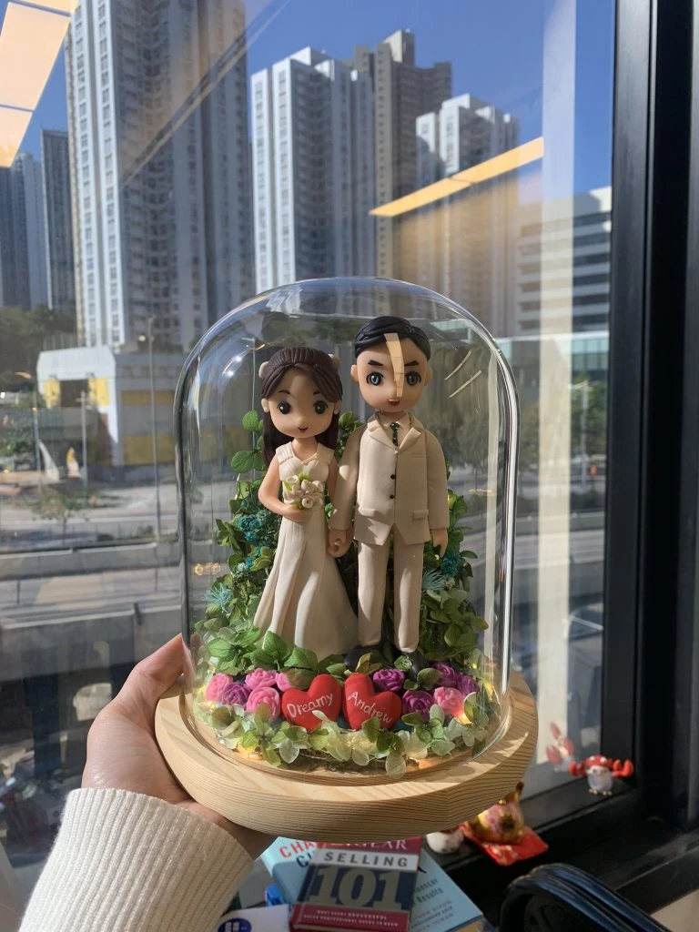 木質底座上的玻璃鐘罩內，Q版新郎新娘並肩站立，新郎著米色西裝、新娘穿白紗捧花，腳邊鋪滿綠葉與紫粉玫瑰花，前方兩顆紅色愛心寫上名字並有暖光燈點綴。