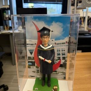 男畢業生主題人像公仔穿深藍學袍戴學士帽,手持文憑站在草地底座與校園背景前,透明展示盒內附金色名牌