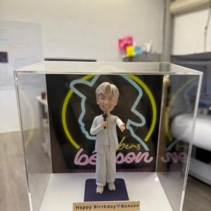 透明防塵盒內的人像公仔:白色系演出造型男子手持麥克風站在紫色地台前,背板為黑底霓虹線條與“benson”字樣;前方小銘牌寫著「Happy Birthday Benson」。
