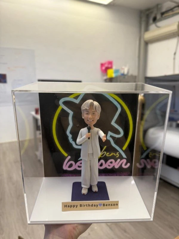 透明防塵盒內的人像公仔：白色系演出造型男子手持麥克風站在紫色地台前，背板為黑底霓虹線條與“benson”字樣；前方小銘牌寫著「Happy Birthday Benson」。