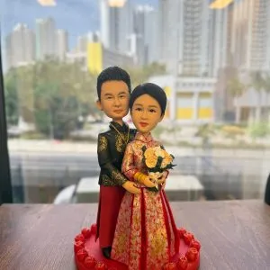 中式婚禮主題公仔：新娘穿紅金龍鳳褂手捧米白玫瑰花束，佩戴金手鐲；新郎著黑金繡紋長衫與紅裙褂，自後環抱新娘。兩人站在紅色愛心底座，周圈環繞立體玫瑰。