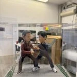 透明展示盒內的雙人公仔：兩名男子在灰色磚道地台上對練站樁交手，左側穿酒紅外套黑褲，右側著深灰運動套裝與白色武術鞋，地台邊緣有草皮與碎花點綴。