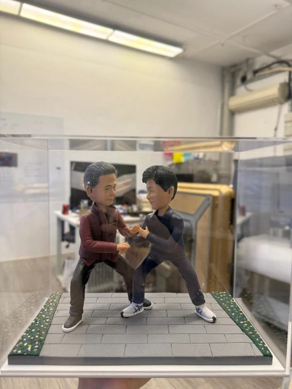 透明展示盒內的雙人公仔：兩名男子在灰色磚道地台上對練站樁交手，左側穿酒紅外套黑褲，右側著深灰運動套裝與白色武術鞋，地台邊緣有草皮與碎花點綴。