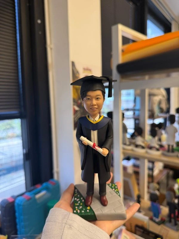 男畢業生人像公仔穿黑色學袍與方帽，手持紅綁帶文憑，站在灰色步道與綠地小花底座上，典禮感十足