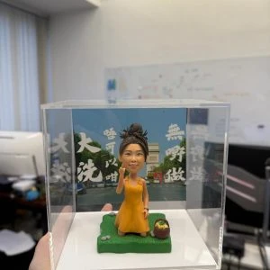 透明展示盒內的人像公仔:綁丸子頭、穿橙黃色洋裝的女孩跪坐在綠色草地地台上,右手做手勢,旁邊有小野餐籃;背板為藍天綠樹與凱旋門街景,覆有白色字樣。