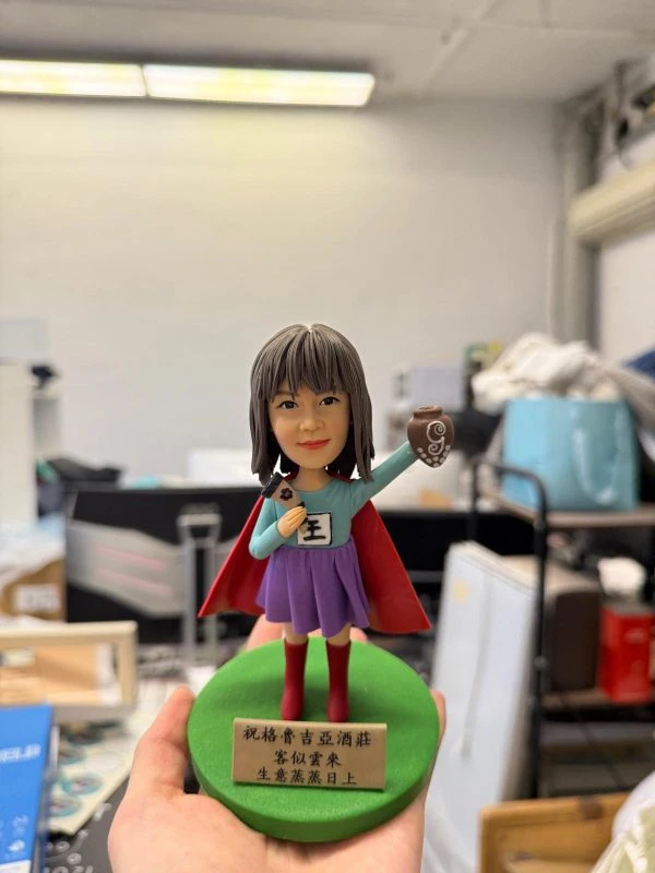 英雄披風女店長公仔高舉咖啡杯與字牌，綠色底座附祝賀銘牌，開幕與業績慶祝客製禮