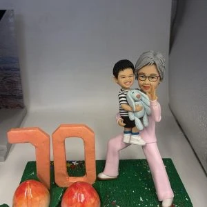 長壽主題公仔擺設:銀髮長輩穿粉色套裝抱著微笑孫子與藍色玩偶熊,綠草地台上有大型「70」與兩顆長壽桃,前方金色銘牌寫上祝福與日期。