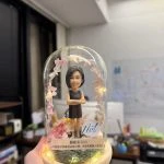 玻璃圓頂罩內的人像公仔：短髮女士穿黑色無袖洋裝、雙手抱臂站立，周圍環繞粉色系乾燥花與小燈串，木質圓形燈座上有苔蘚草地與銘牌，側邊貼有「Neit」標誌。