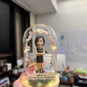 玻璃圓頂罩內的人像公仔:短髮女士穿黑色無袖洋裝、雙手抱臂站立,周圍環繞粉色系乾燥花與小燈串,木質圓形燈座上有苔蘚草地與銘牌,側邊貼有「Neit」標誌。