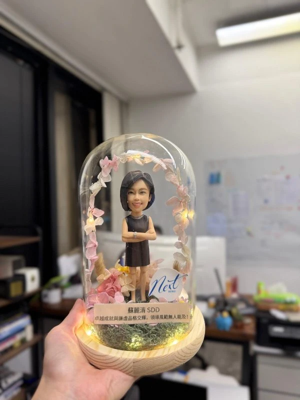 玻璃圓頂罩內的人像公仔：短髮女士穿黑色無袖洋裝、雙手抱臂站立，周圍環繞粉色系乾燥花與小燈串，木質圓形燈座上有苔蘚草地與銘牌，側邊貼有「Neit」標誌。