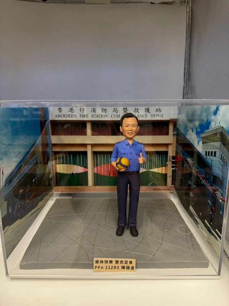 透明展示盒內的人像公仔：身穿藍色制服的男子手抱黃藍排球，站在灰色地台上；背板為「香港仔消防局暨救護站」建築外觀，前方金色銘牌刻有祝福語與編號。