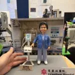 醫護主題人像公仔穿藍色外套與醫護服，站在專科門診背板前，旁邊有血壓監測儀器模型與祝賀銘牌，前方擺放「Grantham Hospital」標牌