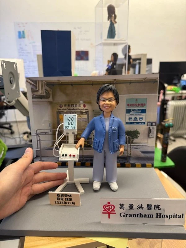 醫護主題人像公仔穿藍色外套與醫護服，站在專科門診背板前，旁邊有血壓監測儀器模型與祝賀銘牌，前方擺放「Grantham Hospital」標牌