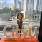 女性人像公仔穿條紋西裝外套站於橙色亮面底座上，前方以立體「CityU」字樣做造型，側前方貼有英中雙語退休祝福銘牌