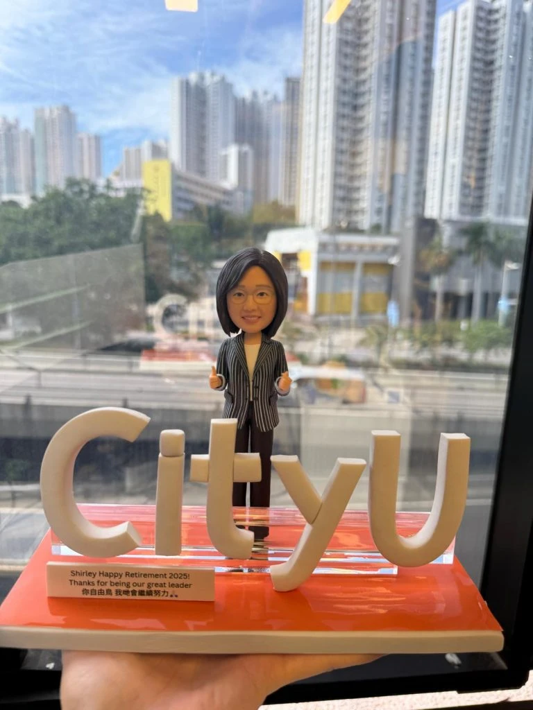 女性人像公仔穿條紋西裝外套站於橙色亮面底座上,前方以立體「CityU」字樣做造型,側前方貼有英中雙語退休祝福銘牌