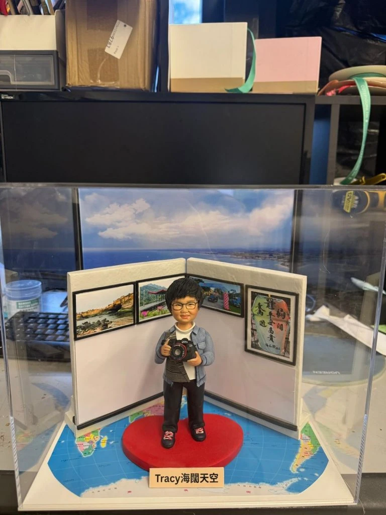 透明展示盒中的攝影主題公仔：短髮戴眼鏡的人物手持相機站在紅色愛心地墊上，背後三折屏風掛有風景照與書法字畫；地台鋪世界地圖，前方銘牌寫「Tracy 海闊天空」。