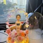 穿和服的人像公仔站在木座上，周圍以粉橘與米白永生花環繞，頭頂有花瓣燈串形成拱門，底部嵌入暖黃燈，旁放透明玻璃罩